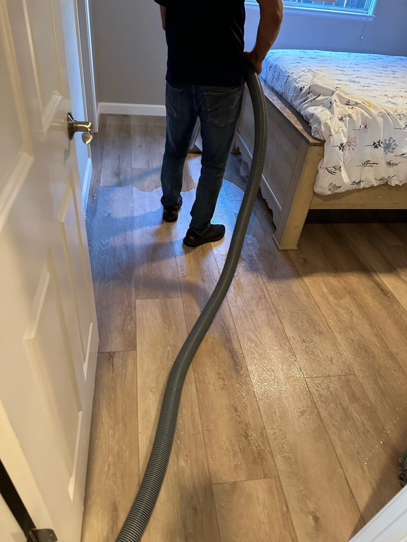 A technician in jeans and a dark shirt pulling an extraction hose across a wet bedroom floor beside a bed.